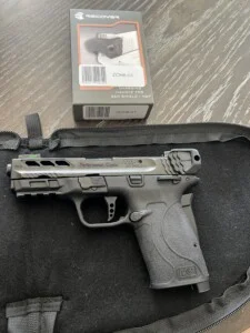 Handgun on a black surface with a nearby box labeled "charging handle for S&W Shield/M&P.