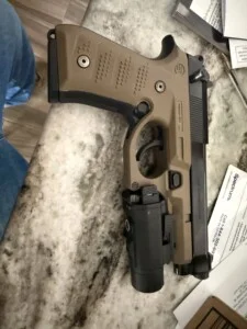 A handgun with an attachment is placed on a marble surface next to papers and a pen.