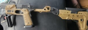 Two tan and black firearms resting on a dark surface, connected by a folding stock.