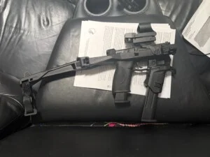 A pistol with a tactical brace and sight attachment rests on documents on a leather seat.