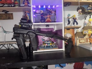 A gun with a stabilizing brace is displayed on a shelf with various anime figurines and collectibles.