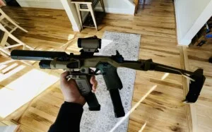 A person holds a tactical firearm with a scope and silencer in a room with wooden floors.